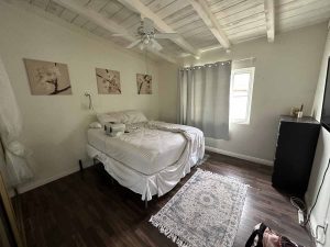 A bedroom with dark wood floors, a rug and bed with white comforter and a painted white wood ceiling with fan.