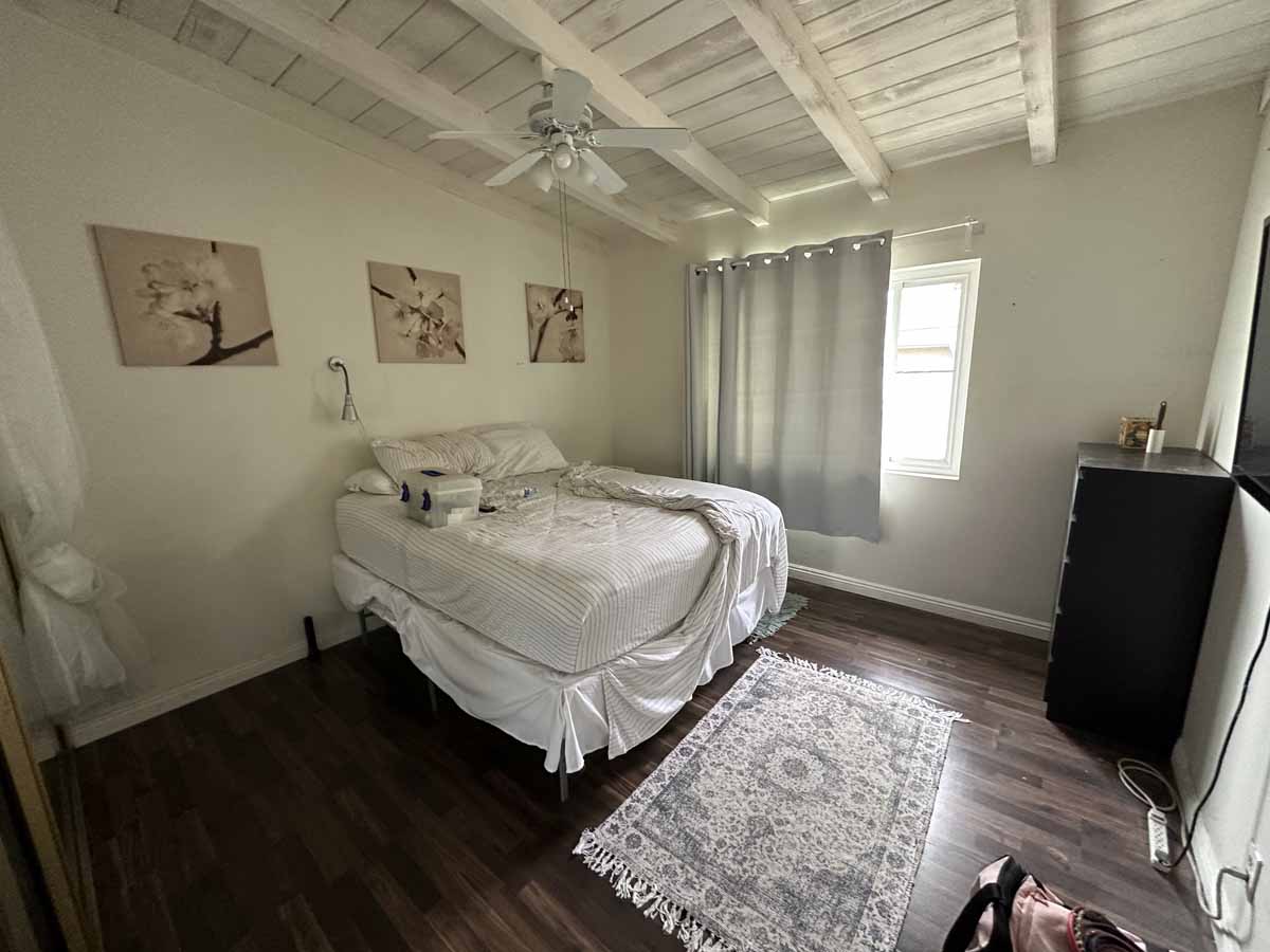 A bedroom with dark wood floors, a rug and bed with white comforter and a painted white wood ceiling with fan.