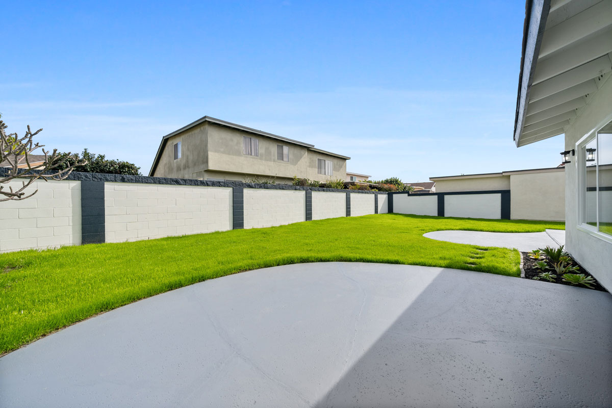 Backyard showing large concrete patio with bright green grass and a concrete block wall.