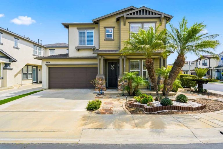 Front of home with beige and brown tones on exterior with palm trees lining the walkway to the front door.