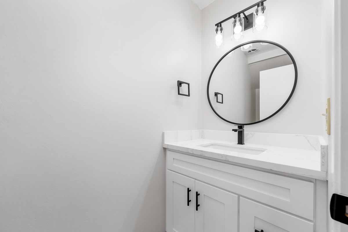 Bright, white, newly renovated bathroom with white stone counter tops on top of a white cabinet, and a black framed circular mirror above the sink.