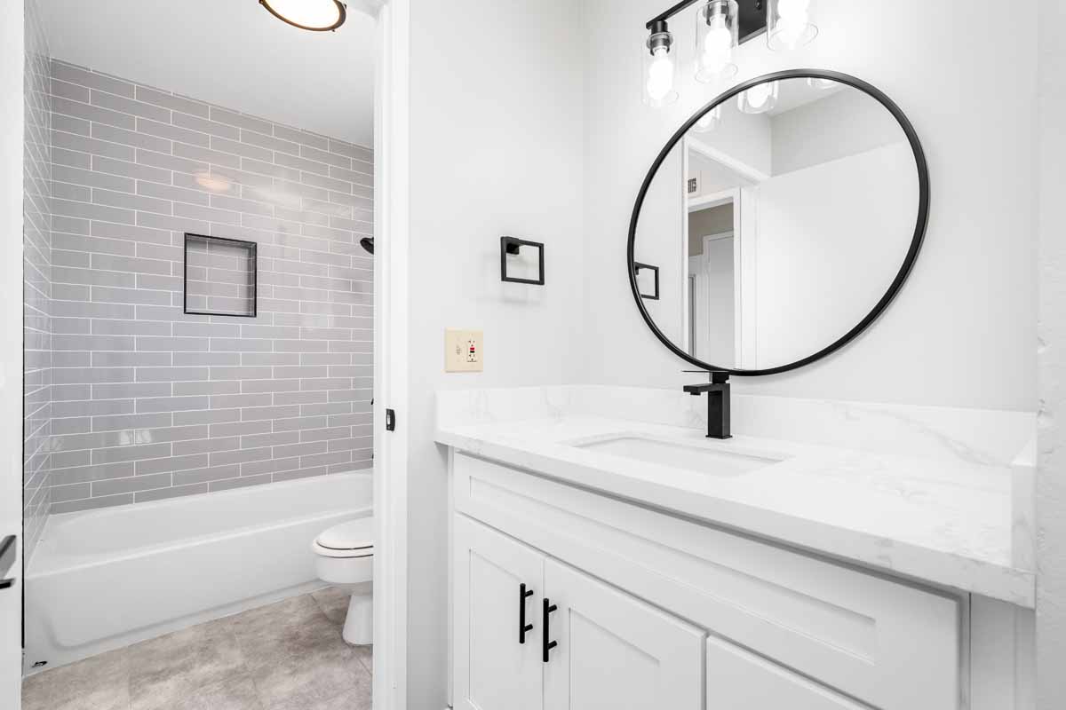 Bright, white, newly renovated bathroom with white stone counter tops on top of a white cabinet, and a black framed circular mirror above the sink.