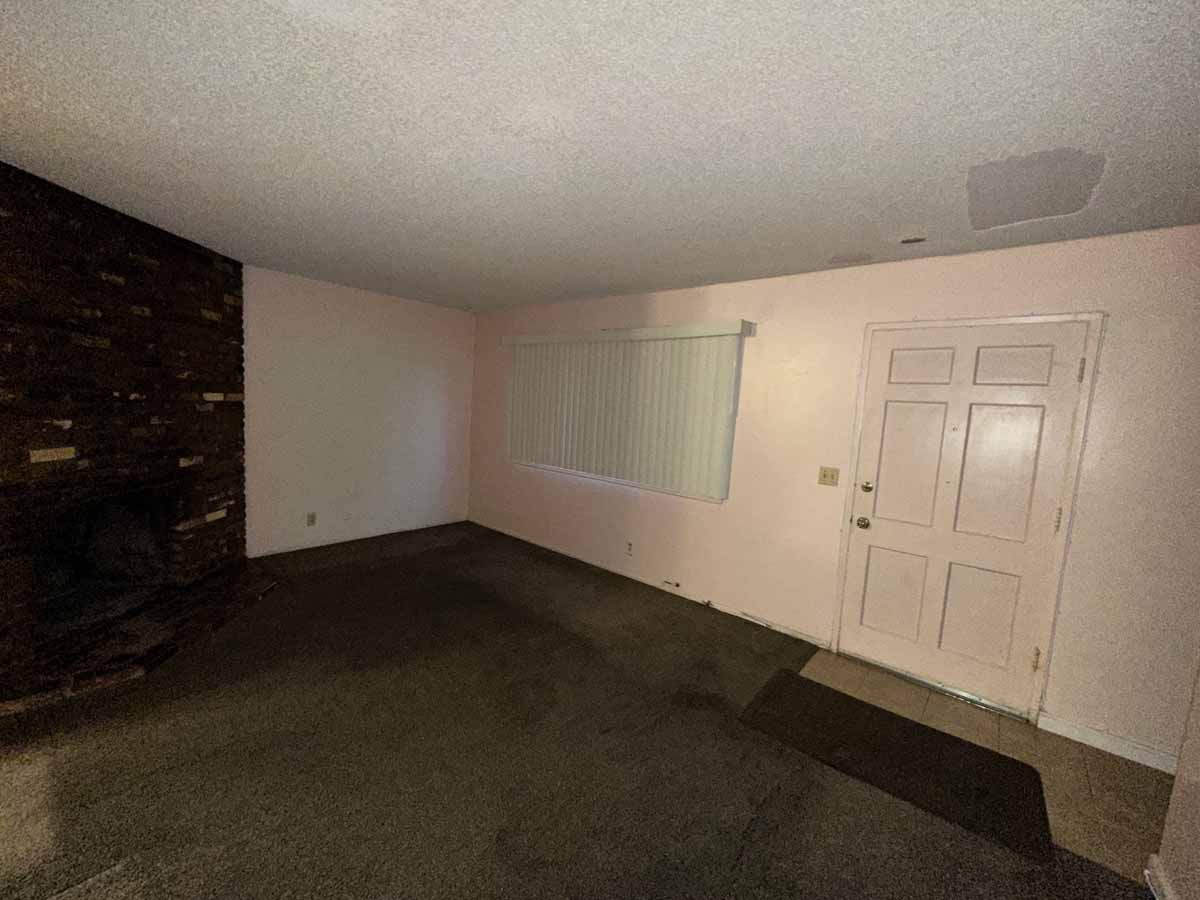 Dark, outdated living room with dark colored carpet, neutral wall color and a dark brink fireplace surround.