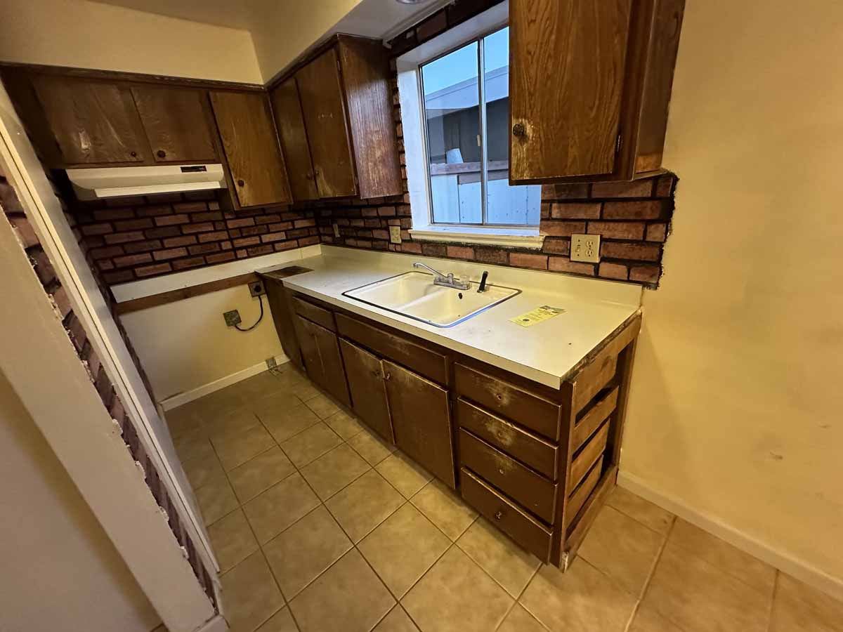 Dark, outdated kitchen with dark wood cabinetry, light colored countertops and beige tile flooring.