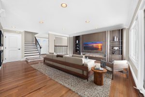 Digitally staged living room with wood floors, a brown couch and stairs in the background.