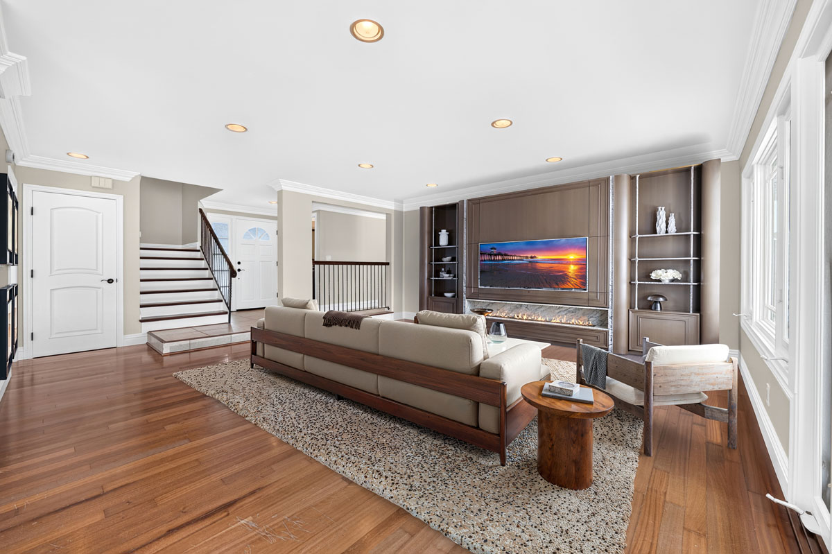 Digitally staged living room with wood floors, a brown couch and stairs in the background.