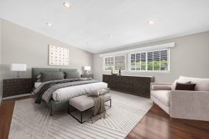 Digitally staged master bedroom with a bed, dresser, chair and large bright window.