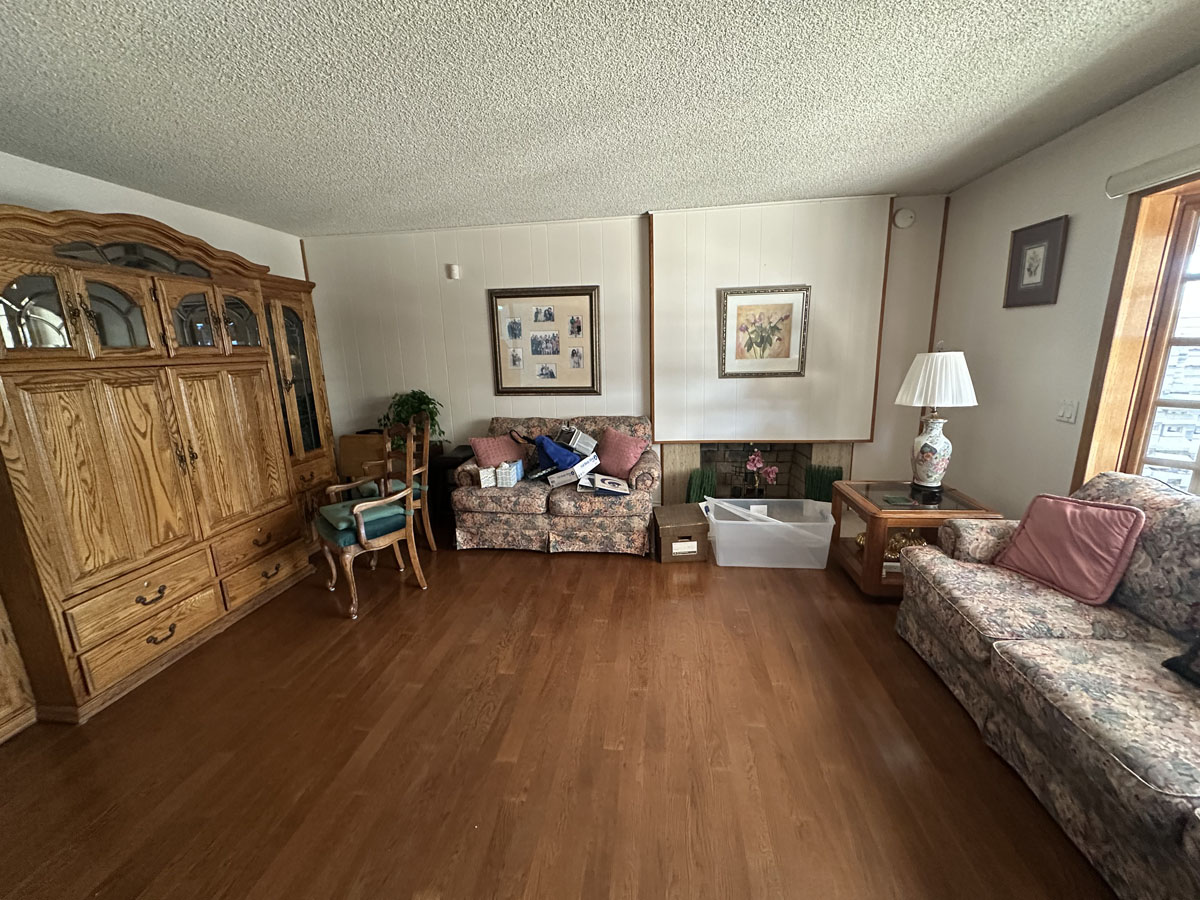 Outdated living room with wood floors and a big window on the back wall.
